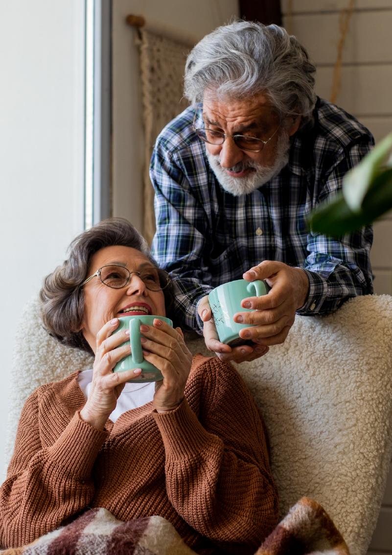 An elderly couple in a cozy kitchen setting, with the man standing behind the woman, embracing her affectionately. The woman is seated and smiling, looking down at a document they are reviewing together. They both wear glasses and casual, comfortable clothing. A pendant light hangs above, and there's a microwave in the background, suggesting a modern home environment.